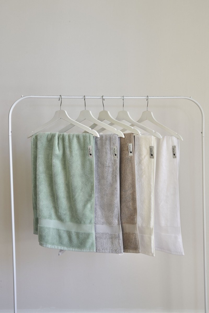 Cinco toallas de bambú en tonos pastel colgadas en perchas de madera blanca sobre fondo neutro, en verde, gris, marrón, crema y