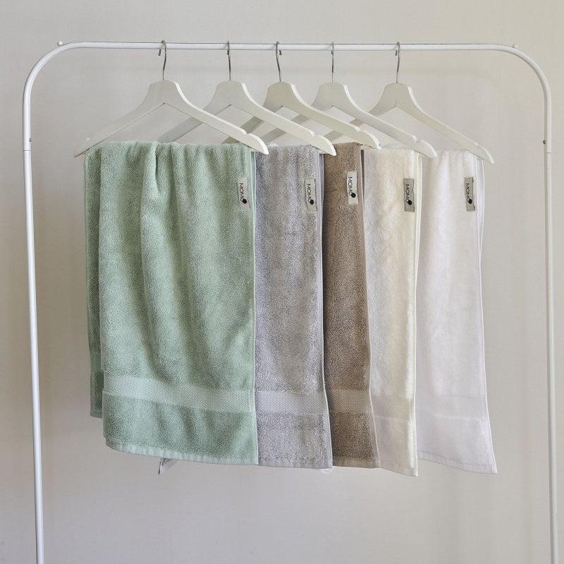 Cinco toallas de bambú en tonos pastel colgadas en perchas de madera blanca sobre fondo neutro, en verde, gris, marrón, crema y