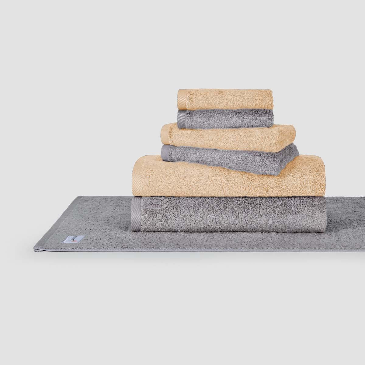 Juego de toallas apiladas beige y gris Momo Cotton mostrado sobre alfombrilla de baño gris en fondo blanco