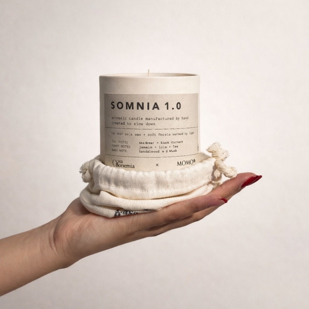 Mano sosteniendo vela aromática Somnia 1.0 color crema envuelta en tela de algodón natural sobre fondo blanco