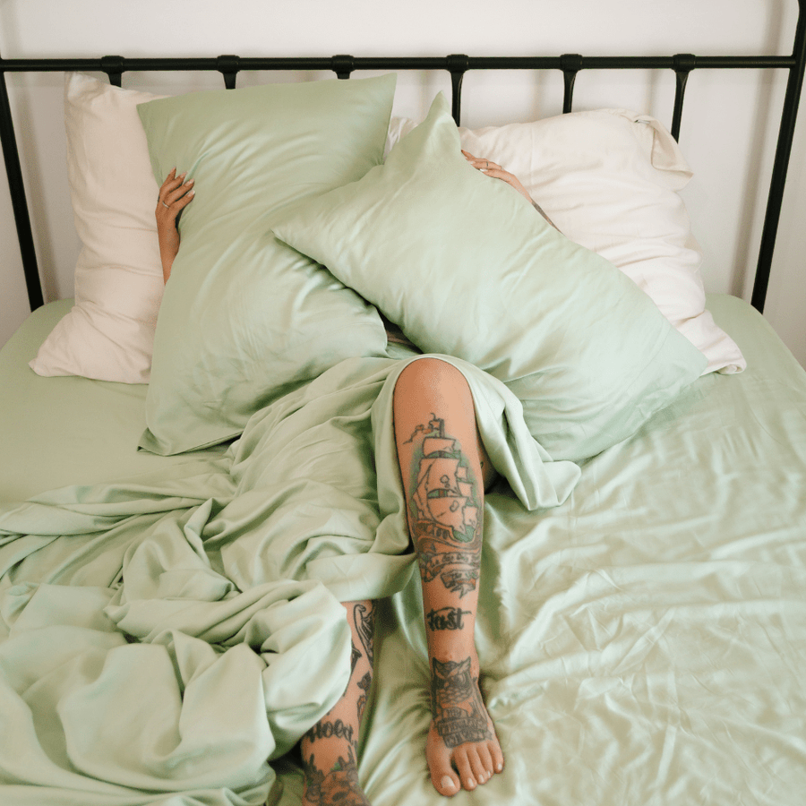 Persona acostada en la cama con juego de ropa de cama de bambú verde y blanco con tatuajes visibles en las piernas