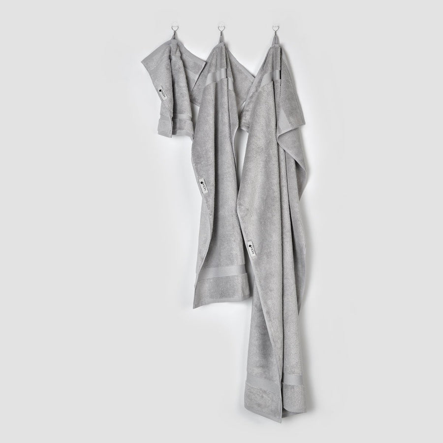 Tres toallas de bambú gris colgadas en ganchos sobre fondo blanco - toallas de cara, mano y baño de la colección Alba