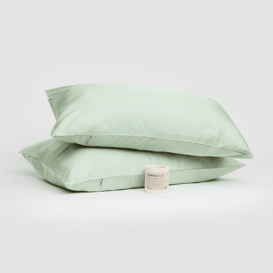 Par de fundas de almohada de algodón verde salvia apiladas con vela Somnia 1.0 sobre fondo blanco