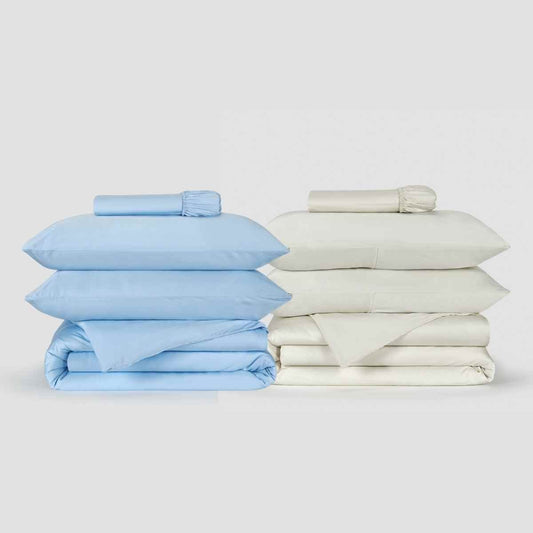 Dos juegos de funda nórdica Momo Cotton lado a lado, uno azul claro y otro crema, con sábanas dobladas y ropa de cama enrollada