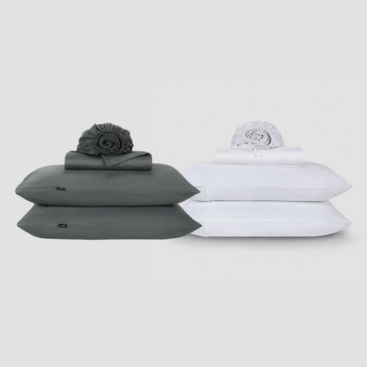 Dos juegos de sábanas Momo Cotton lado a lado, uno gris carbón y uno blanco, cada uno apilado con almohadas y enrollado